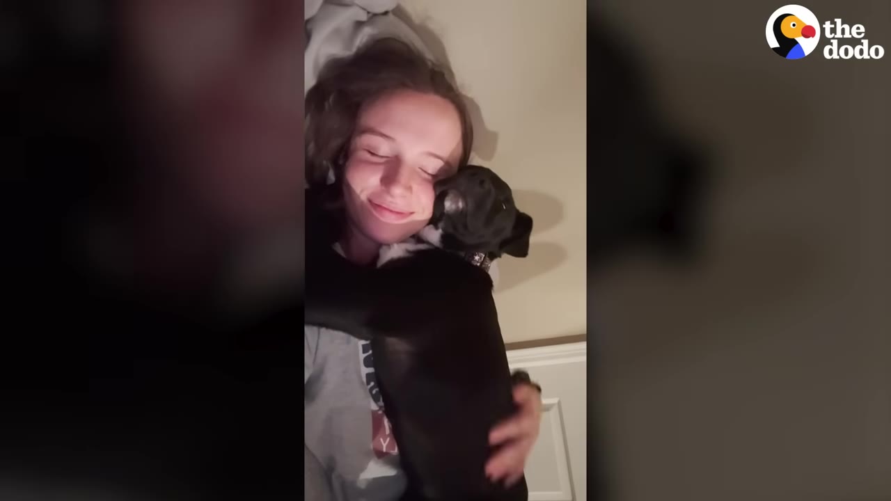 A smiling woman lying down and hugging a small black dog close to her chest.