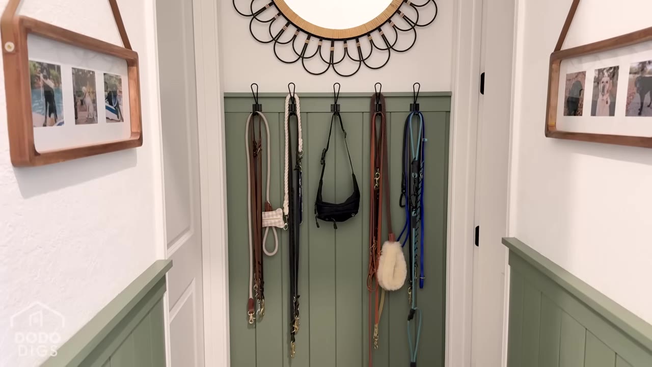 An organized entryway wall with green wainscoting, featuring five black hooks hanging various dog leashes, a black bag, and a fluffy white dog accessory, beneath a round mirror with a decorative black metal frame.