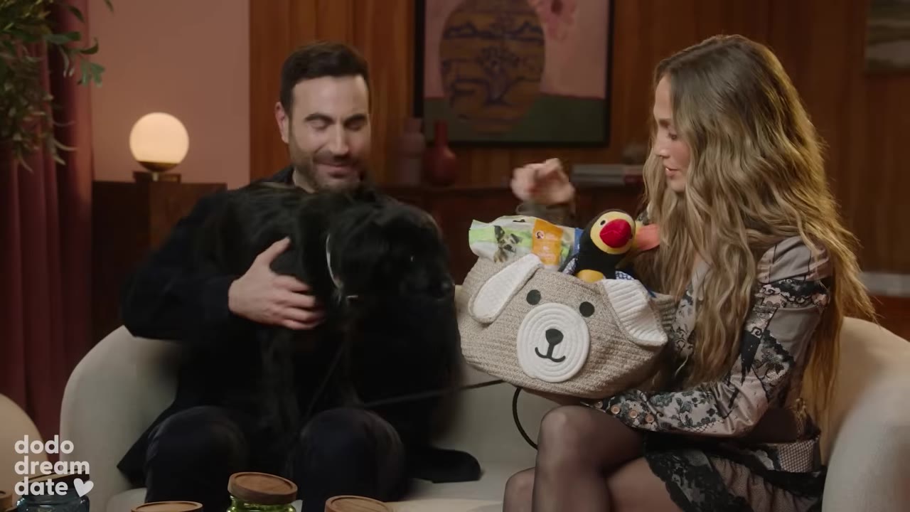 A woman holds a dog-themed gift basket filled with toys and treats next to a man holding a small black dog.