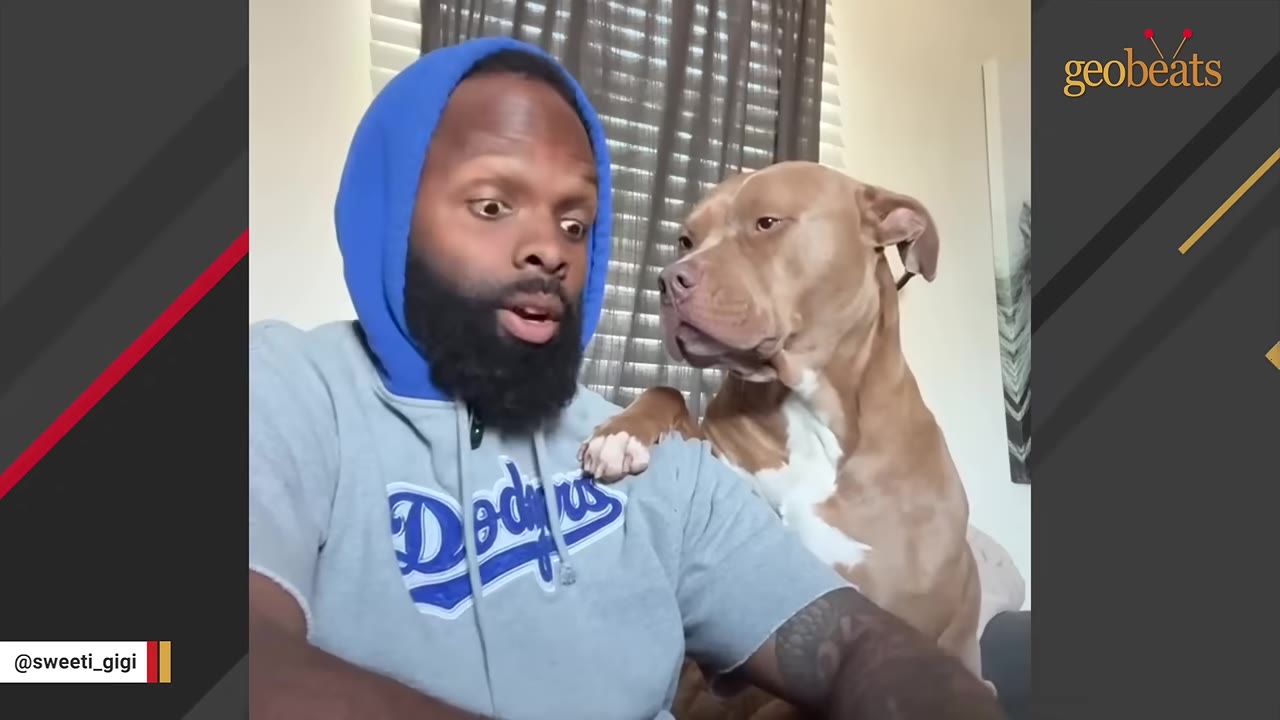 A man in a blue hooded sweatshirt and grey Dodgers t-shirt looks at his light brown pit bull dog, who has placed its paw on his shoulder.