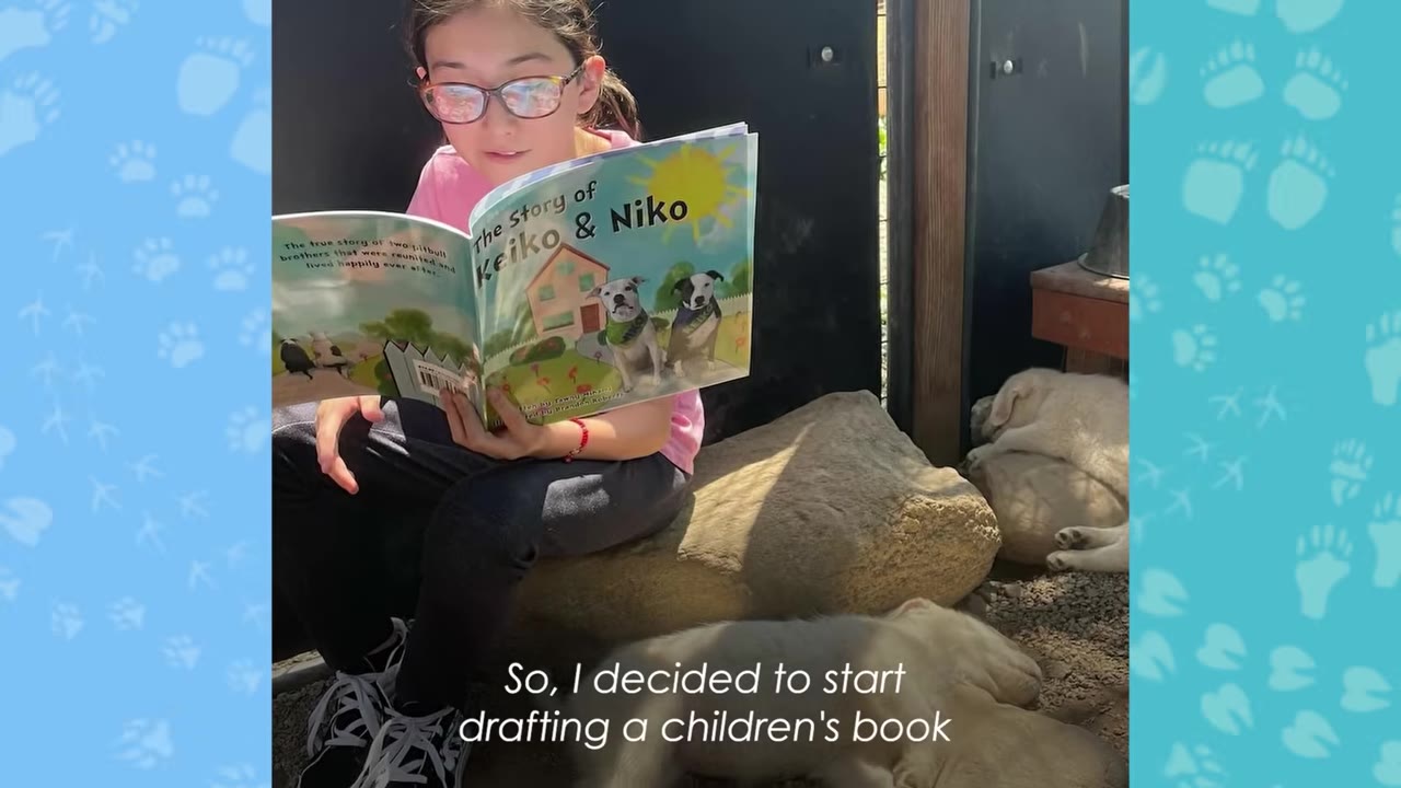 A young girl sitting outdoors reads an open children's book titled 'The Story of Keiko & Niko' which features illustrations of two dogs on the cover.