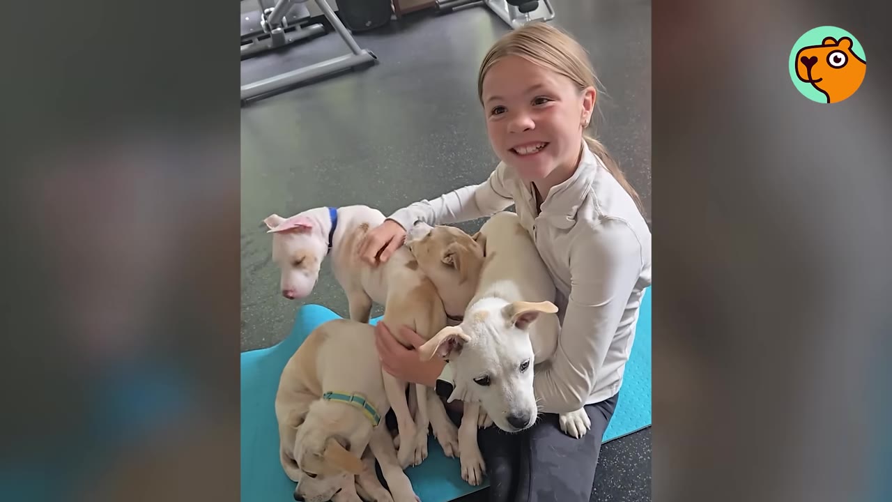 A smiling young girl sitting on a blue yoga mat, surrounded by and cuddling four light-colored puppies.