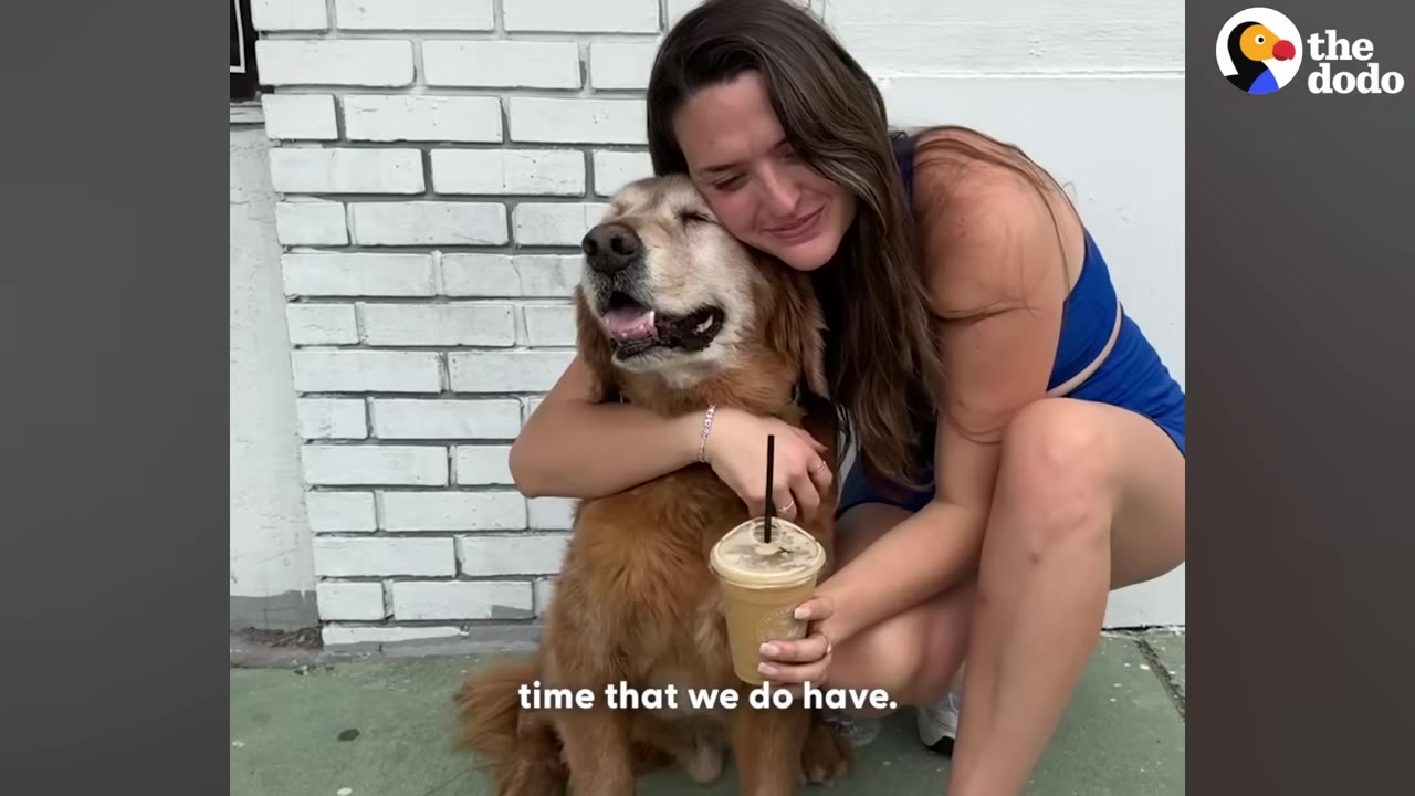A woman in a blue outfit hugs a golden retriever dog tightly while smiling, holding a plastic cup with a straw.