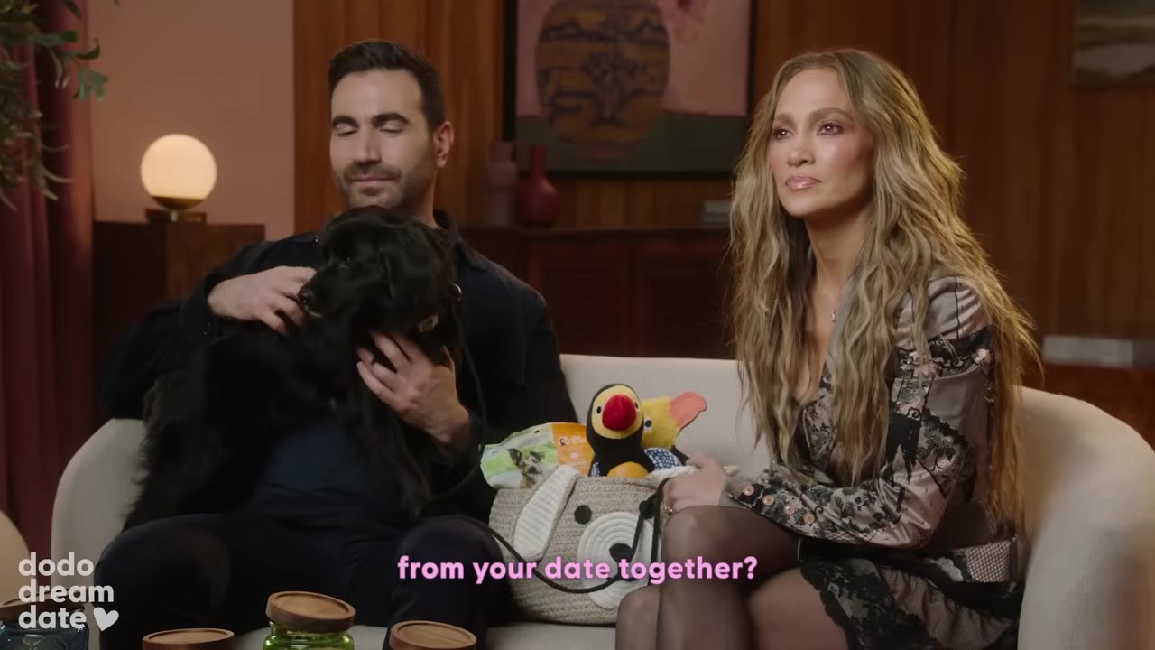 Brett Goldstein sits on a couch holding a small black dog named Regina next to Jennifer Lopez.