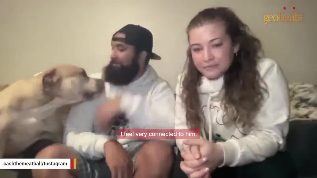 A tan dog nuzzles a bearded man wearing a black cap, while a woman with long wavy hair sits next to them on a couch.