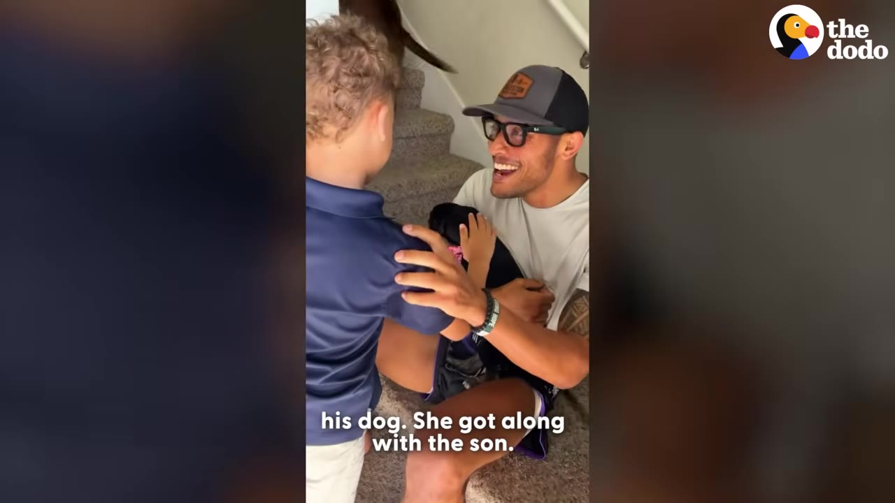 A smiling man wearing a baseball cap and glasses holds a small black puppy while a young boy stands next to him on the stairs.