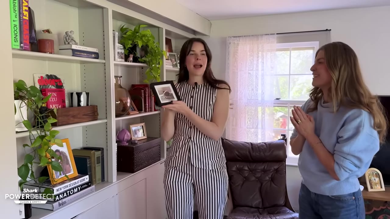 A woman in a striped jumpsuit holds up a small framed photograph of a dog while standing in a living room next to another woman.