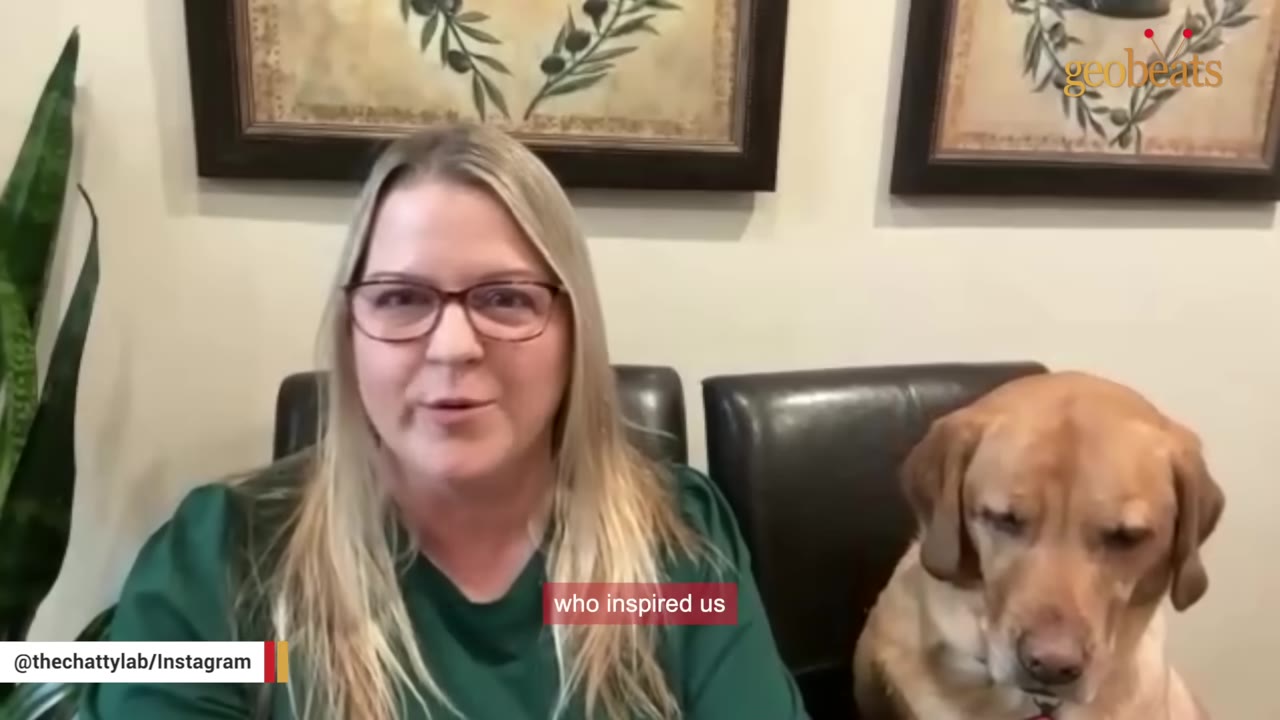 A woman with blonde hair and glasses sits next to a yellow Labrador retriever while speaking.