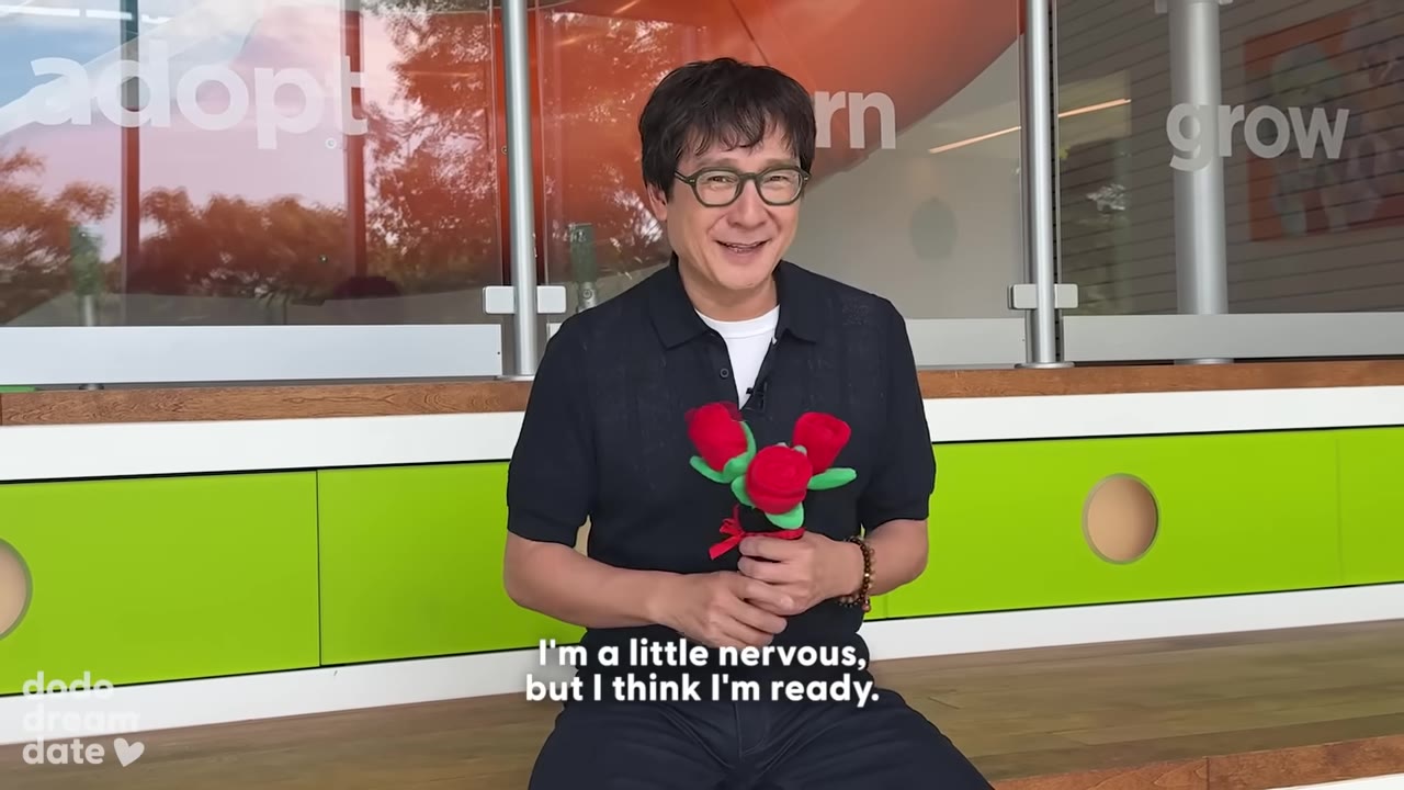 Ke Huy Quan wearing glasses and a black polo shirt, smiling while holding a small bouquet of plush red roses.