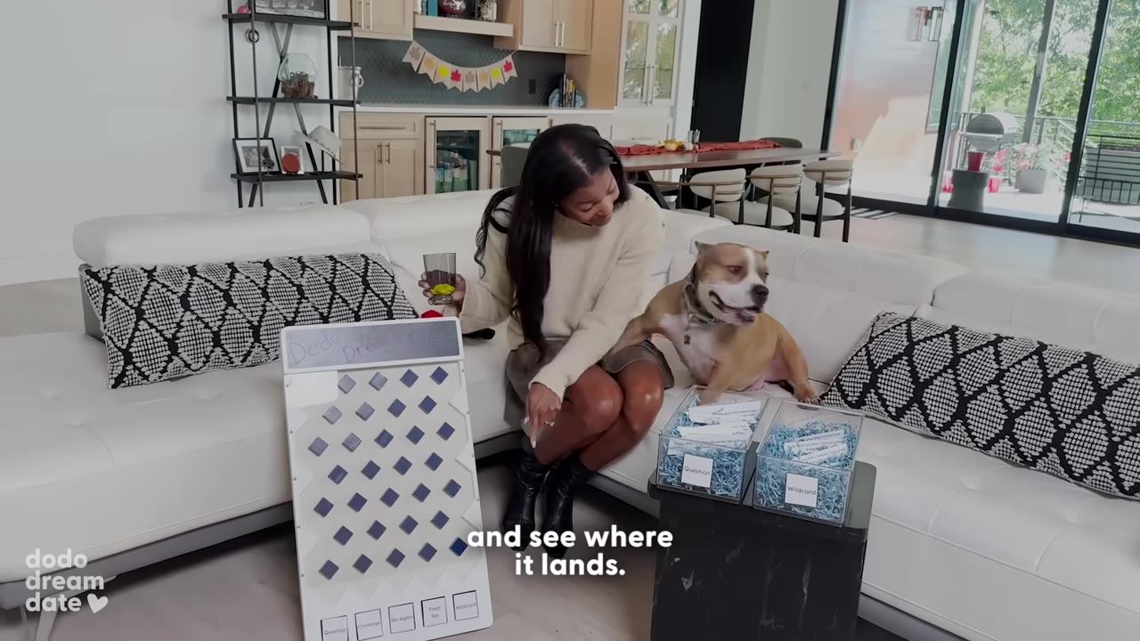 A woman sits on a white couch next to a brown and white dog, pointing at a white Plinko board on the floor in front of her.