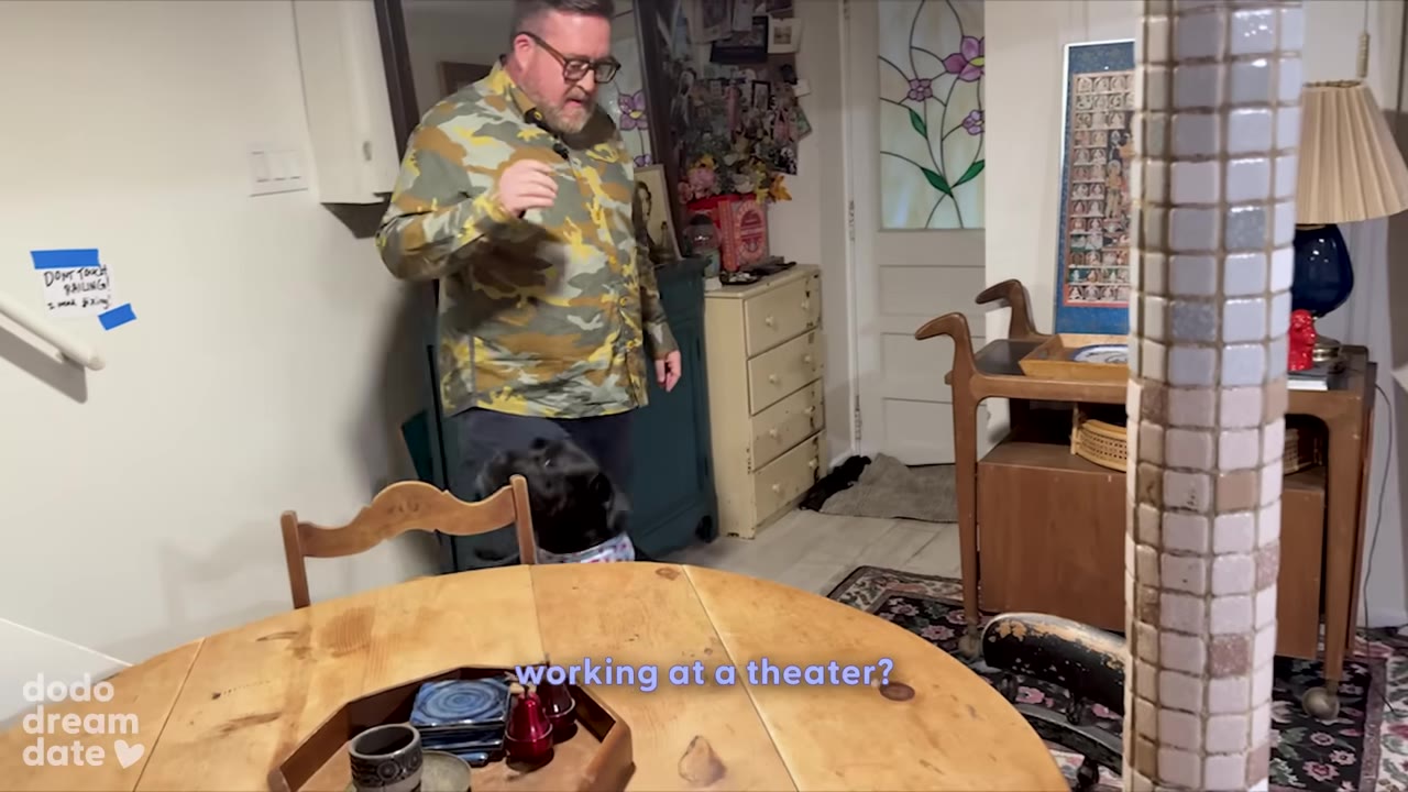 A man in a camouflage shirt stands in a dining room next to a black dog, with a subtitle that reads 'working at a theater?'.