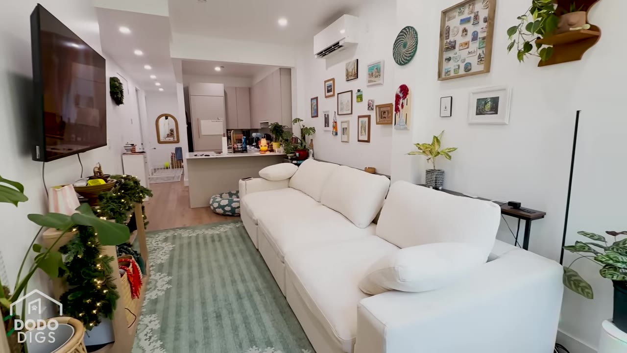 A wide shot of a narrow Brooklyn apartment living room featuring a long white sectional sofa, a green patterned rug, and a gallery wall with framed art.