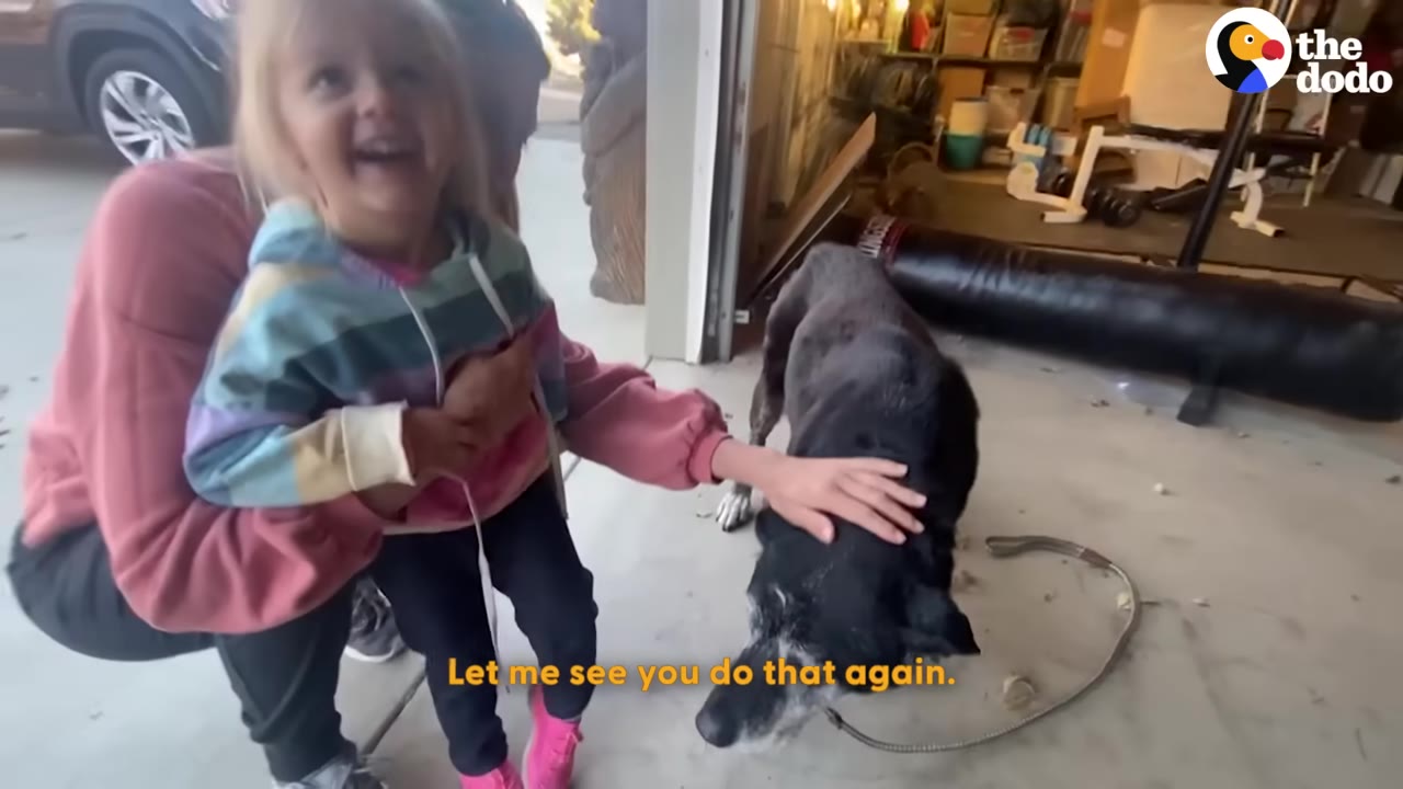 A young girl in a colorful hoodie smiles as a woman kneels beside her, gently petting a black dog named Henry on the head.
