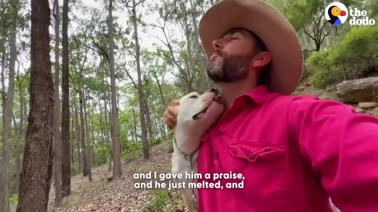 A white dog affectionately nuzzles the neck of a man wearing a cowboy hat and a bright pink shirt in a wooded area.