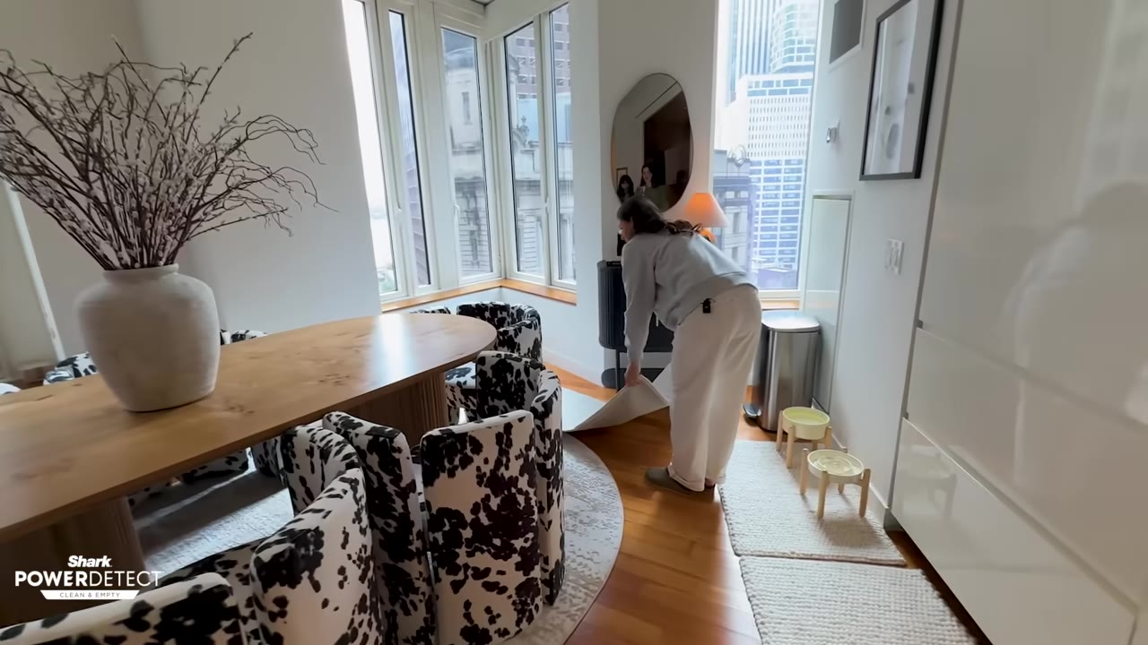 A woman in a grey sweatshirt and white pants lays down a white runner on the hardwood floor of an apartment dining area.