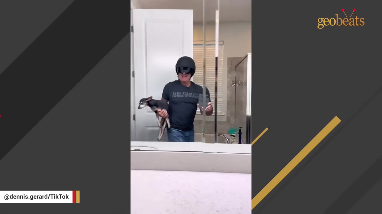 A man wearing a black helmet and black t-shirt holds a small black and tan dog in his arm while taking a mirror selfie in a bathroom.