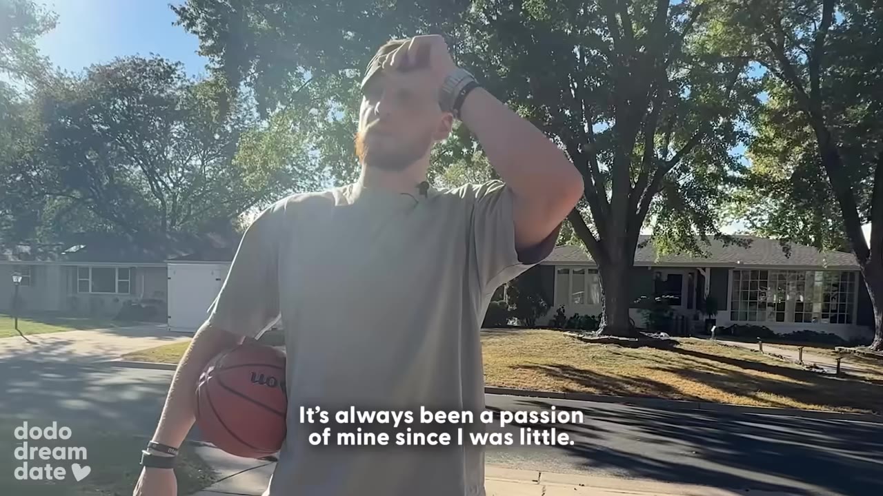 A man holding a basketball under his arm stands on a sunny street, looking off-camera with one hand on his forehead.