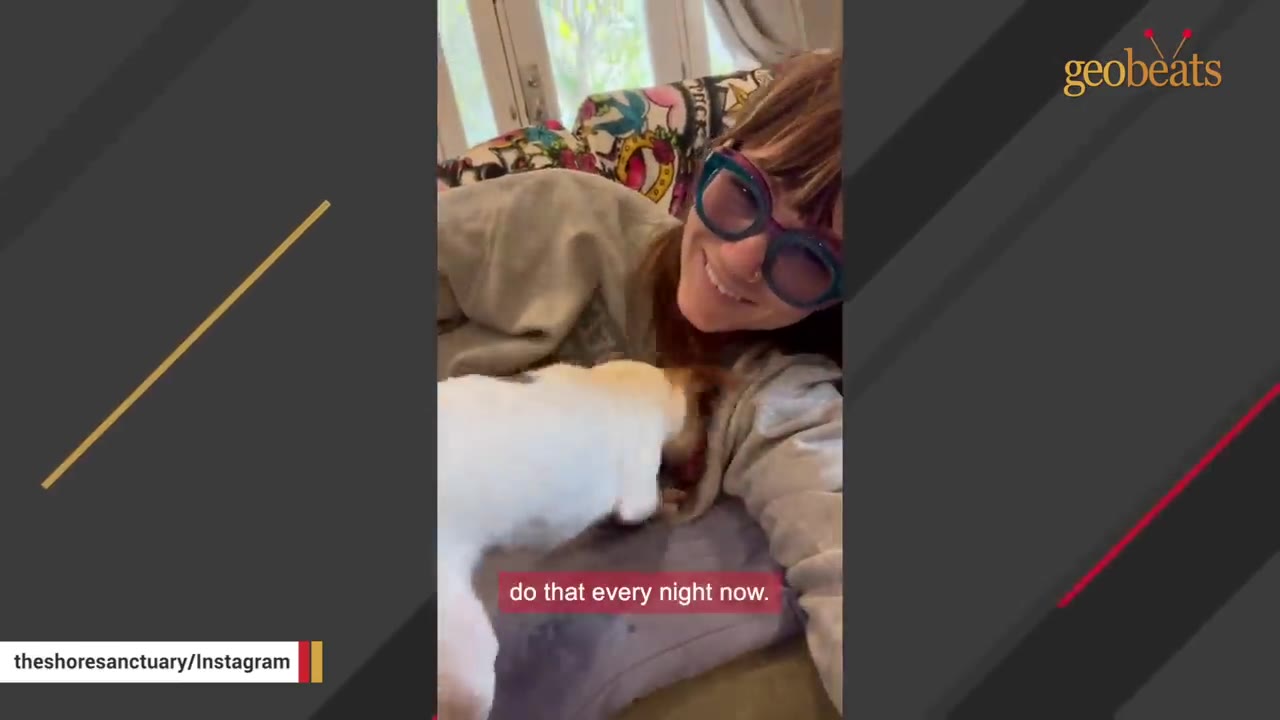 A woman wearing glasses smiles while lying on a sofa as a small white and brown dog cuddles close to her chest.