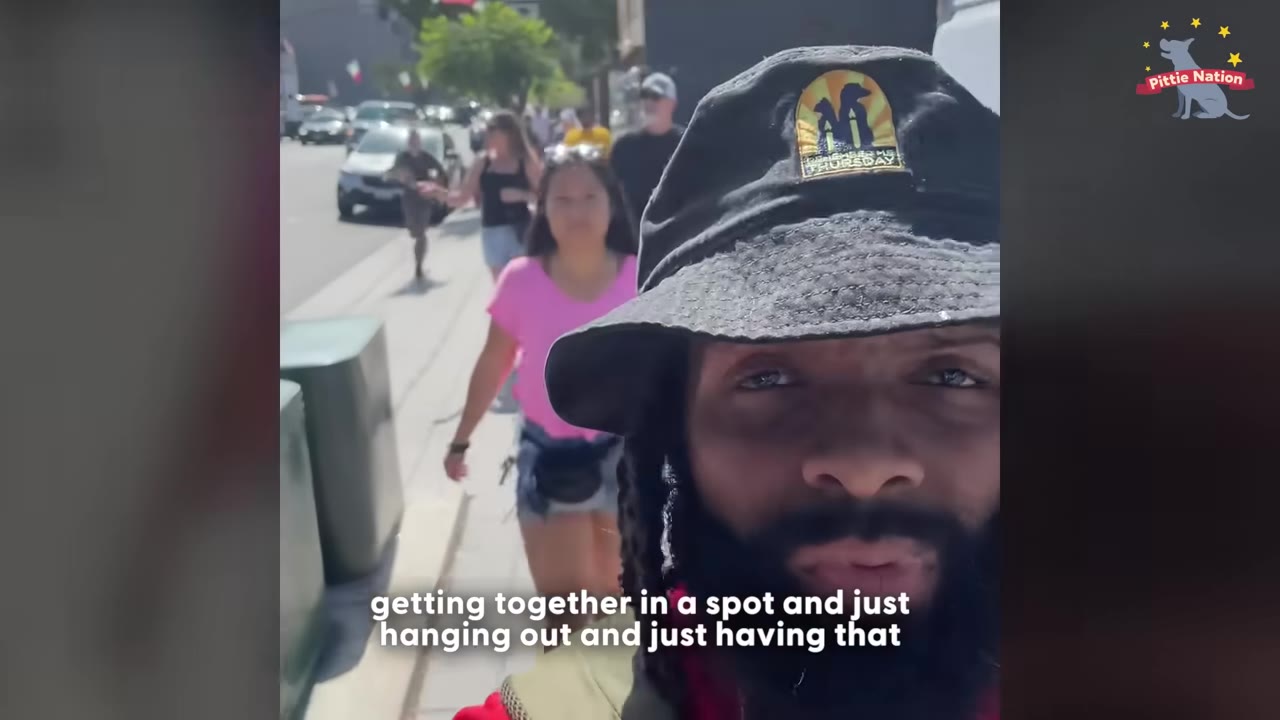 A close-up selfie video frame of a man wearing a black bucket hat and beard, with a woman in a pink shirt walking behind him on a city sidewalk.
