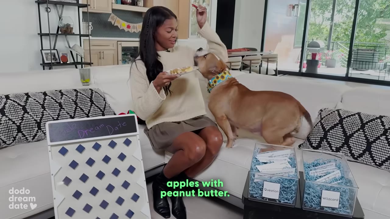 A woman sitting on a white couch feeds a brown dog a treat from a plate, with the caption 'apples with peanut butter.' on the screen.