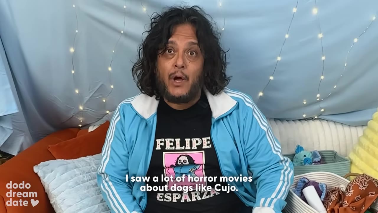 A man with long curly dark hair and a beard, wearing a blue track jacket over a black t-shirt with his name 'Felipe' on it, looks directly at the camera with a wide-eyed, surprised expression.