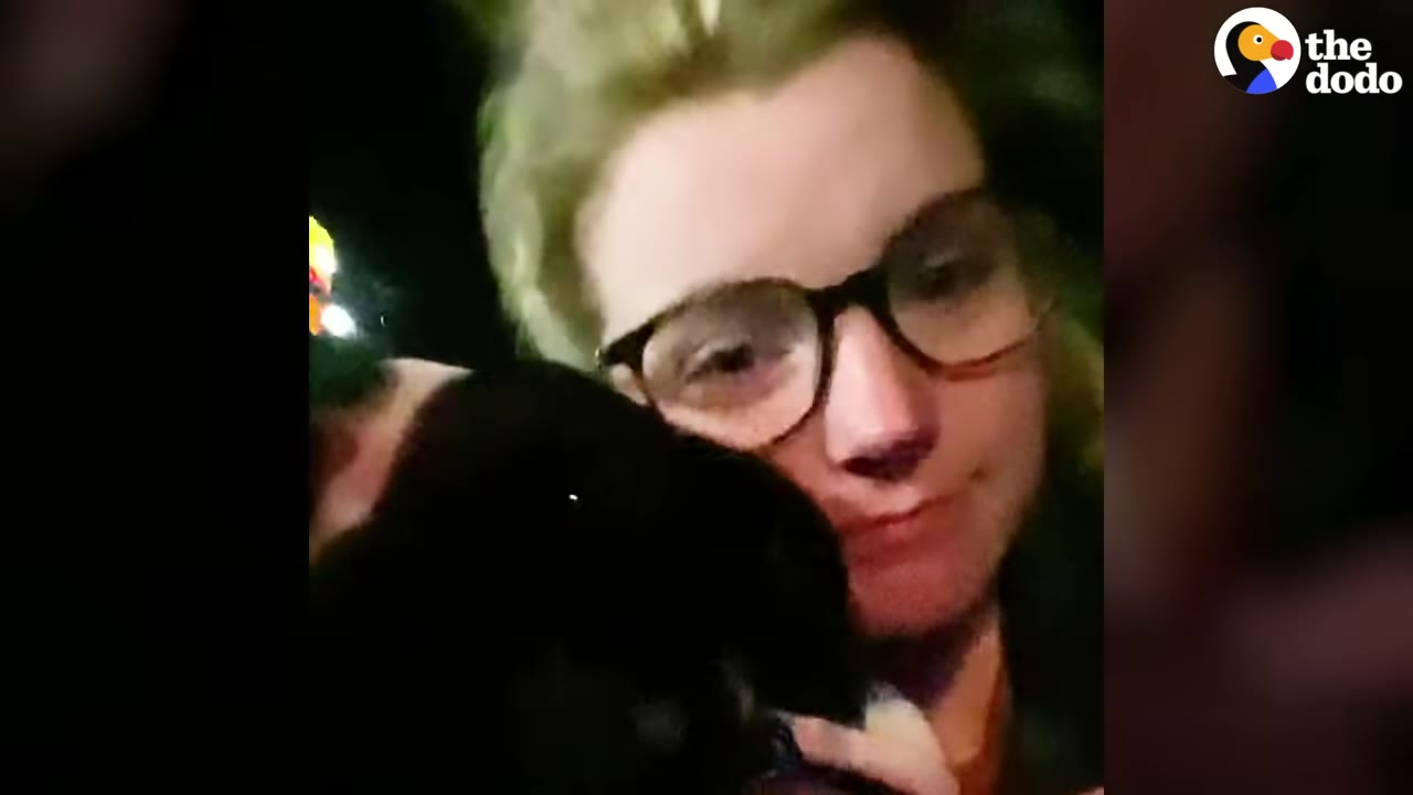 A close-up of a woman wearing glasses holding a black dog close to her face with a bittersweet expression.