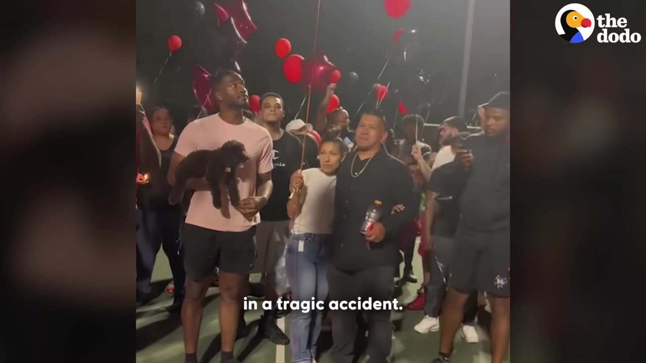 A group of people holding red and black balloons at an outdoor night vigil, with one man holding a small black dog.