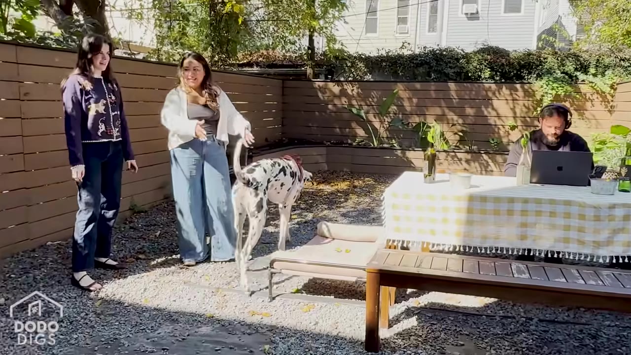 Two women stand in a gravel-covered backyard with a wooden fence, while a man sits at a table with a laptop and a spotted dog walks nearby.