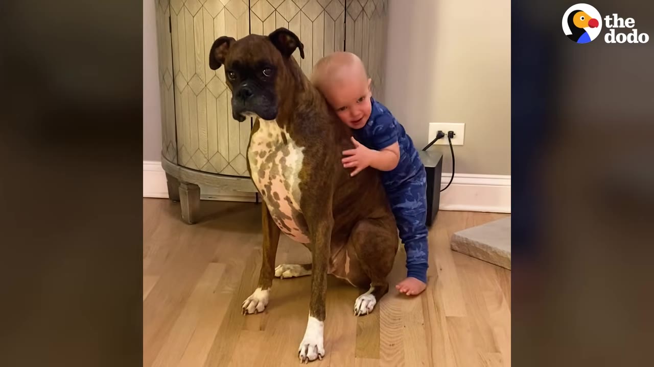 A young toddler in blue pajamas smiles as he hugs a brindle boxer dog from behind.