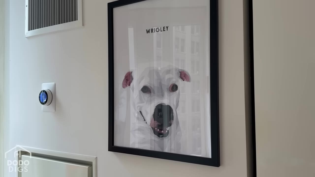 A framed illustration of a white dog named Wrigley hanging on a white wall next to a thermostat.