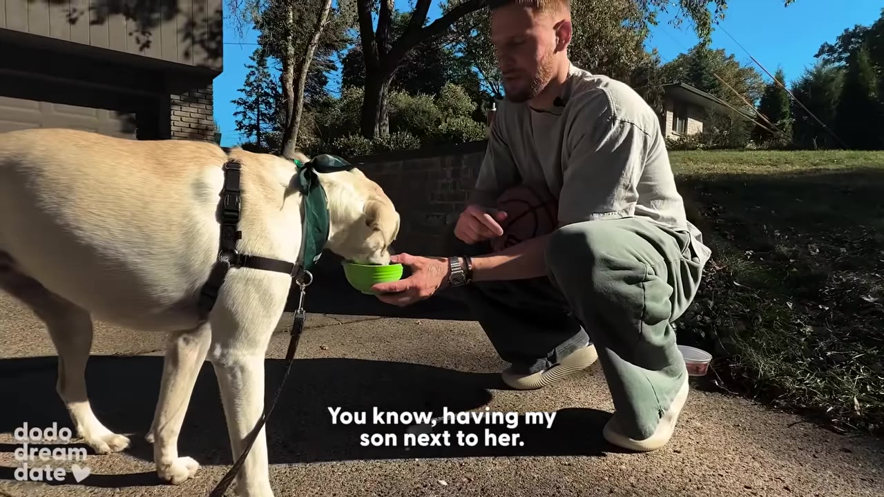 A man crouches down on a paved driveway, holding a small green bowl for a light-colored dog wearing a green bandana to eat from, with subtitles reading 'You know, having my son next to her.'