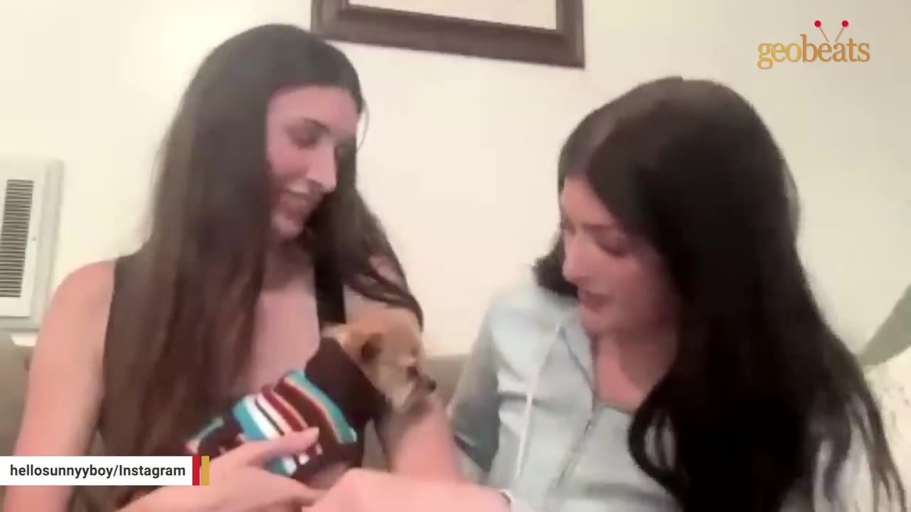 Two women sitting on a couch, smiling and looking down at a small brown dog wearing a striped sweater.