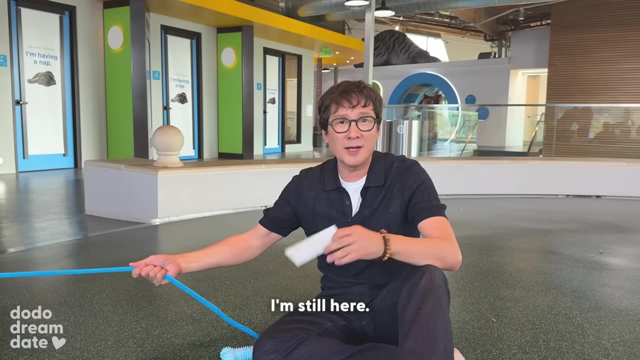 Ke Huy Quan sitting on the floor, holding a blue leash and a white card, looking directly at the camera with a reflective expression, with the subtitle 'I'm still here.'