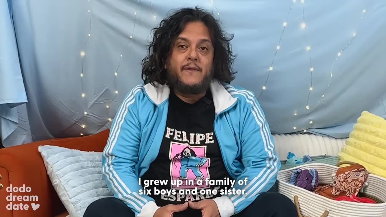 A man with long dark hair and a beard, wearing a blue track jacket over a black graphic t-shirt, sits in front of a blue backdrop with fairy lights.