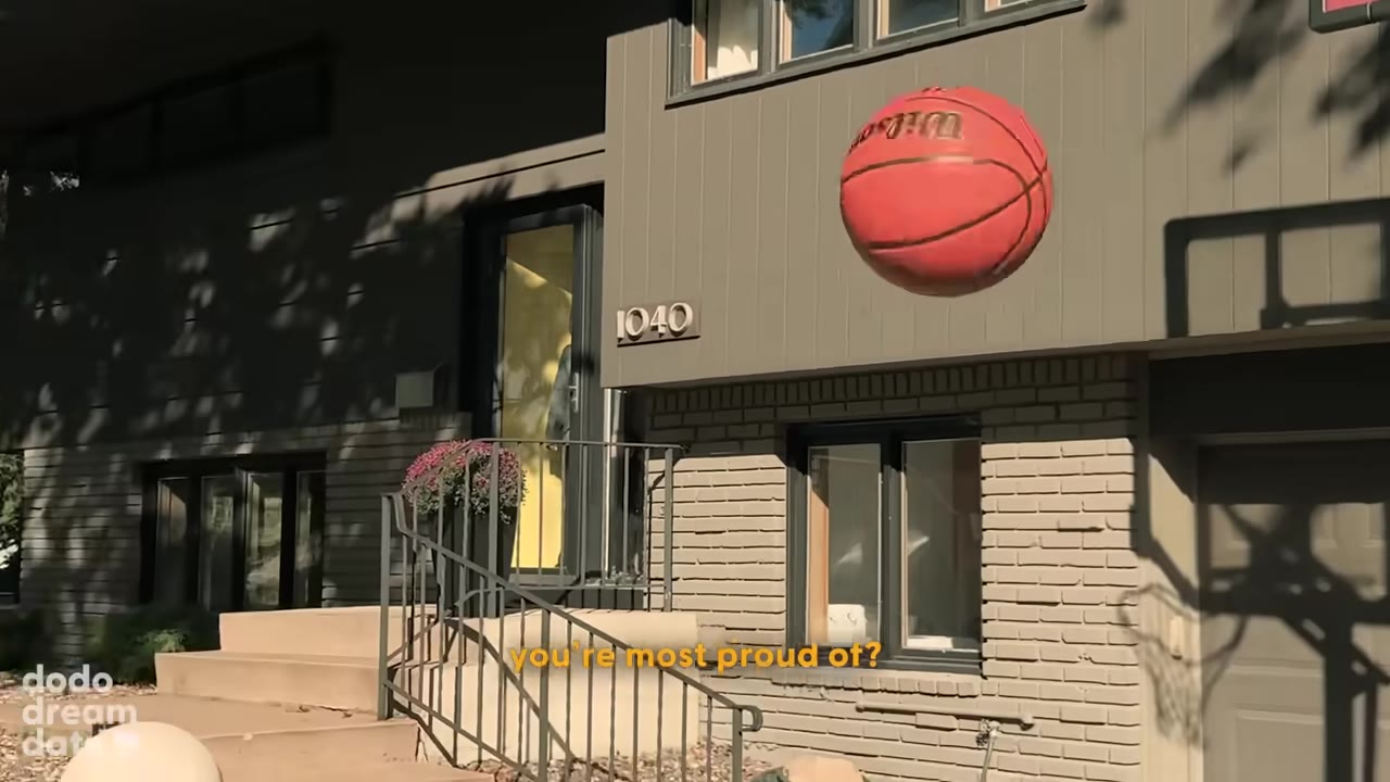 A red basketball flies through the air in front of a modern grey house with a shadow of a basketball hoop on the garage door.