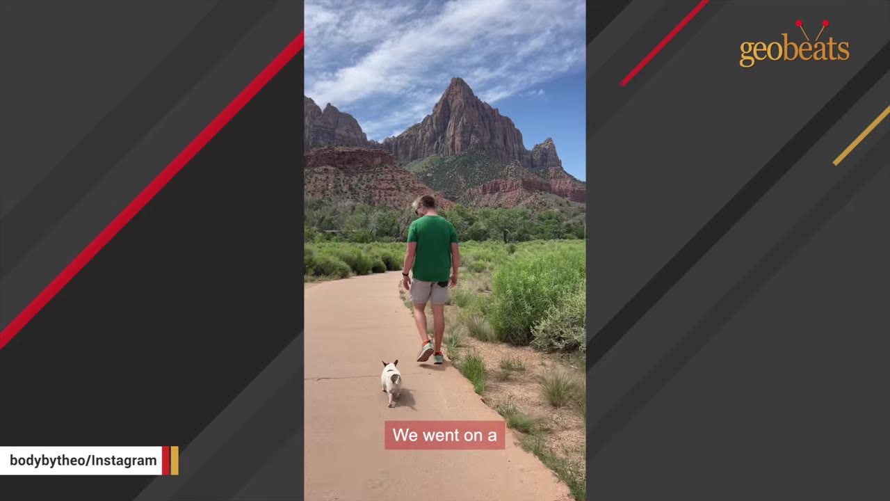 A man in a green shirt walks a small white dog on a paved path with a large red rock mountain in the background.