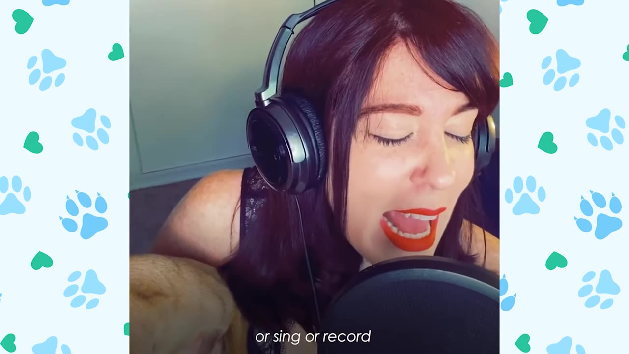 A close-up of a woman with red hair wearing headphones and singing into a studio microphone, with the top of a dog's head visible in the lower left corner.
