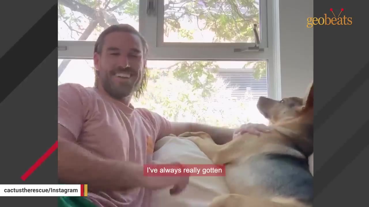 A smiling man sits on a couch next to a German Shepherd mix dog who is lying down and looking up at him.