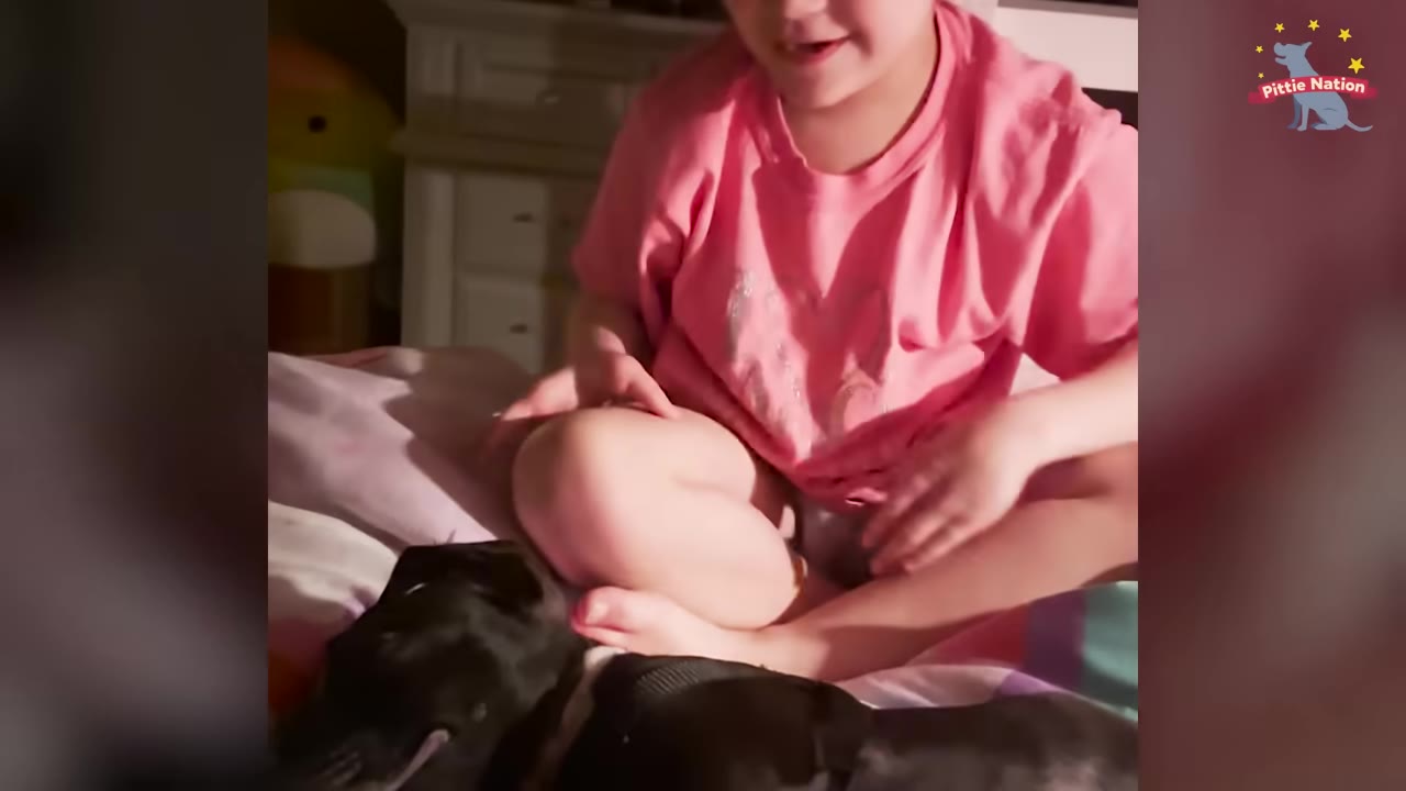 A young girl in a pink t-shirt sits cross-legged on a bed next to a black dog lying down beside her.