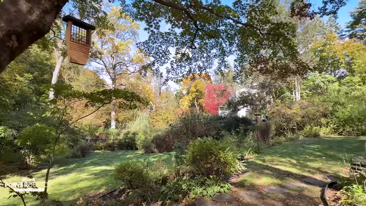 A lush, expansive backyard garden with tiered lawns, dense green shrubbery, and colorful autumn trees under a blue sky.