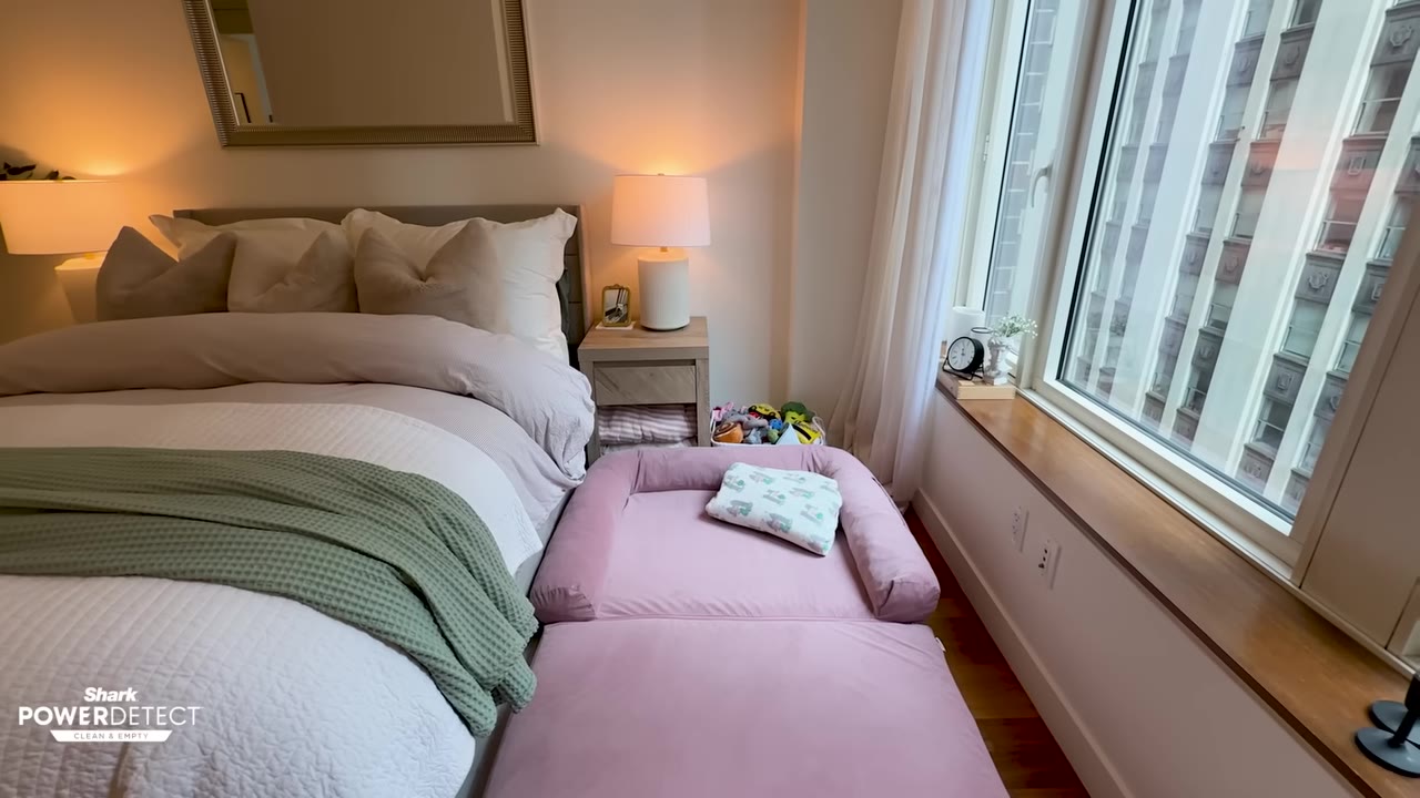 A neatly made bed with white and green bedding next to a pink dog bed in a modern bedroom with a large window.
