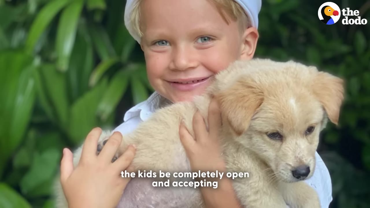 A smiling young boy wearing a white hat holds a fluffy, light-colored puppy in his arms against a background of green leaves.