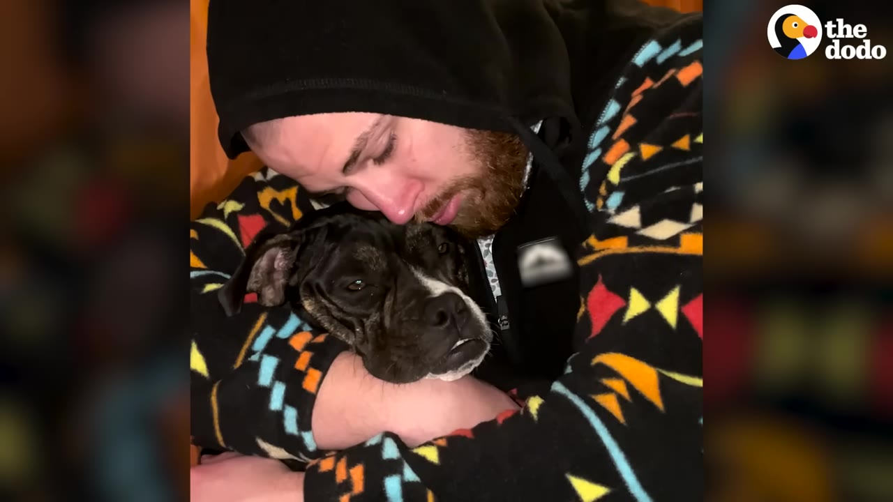 A man wearing a black hood and a colorful patterned fleece jacket hugs a black and white dog with a prominent underbite close to his chest.
