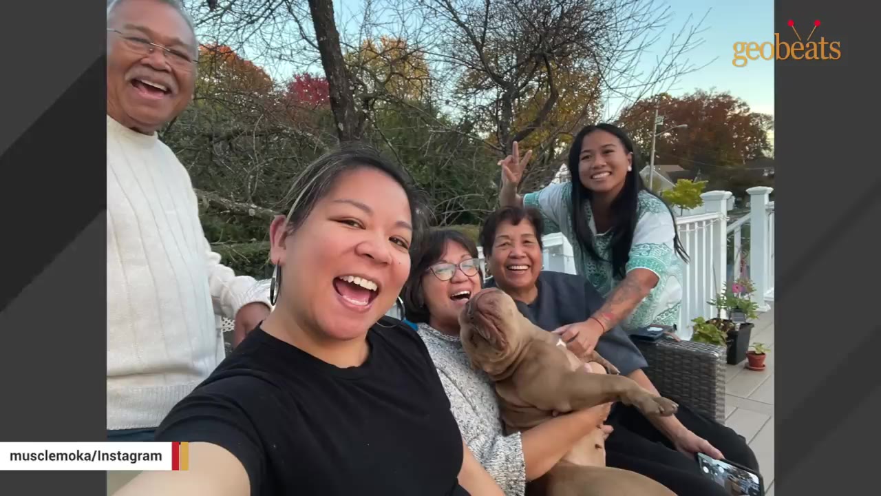 A smiling family poses for a outdoor selfie together, with one woman holding a brown dog in her lap.