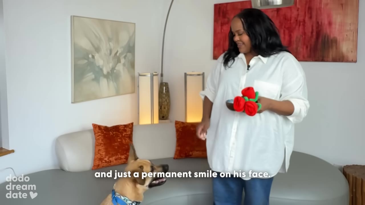 A woman in a white shirt smiles down at a tan rescue dog looking up at her with an open mouth, in a living room setting.