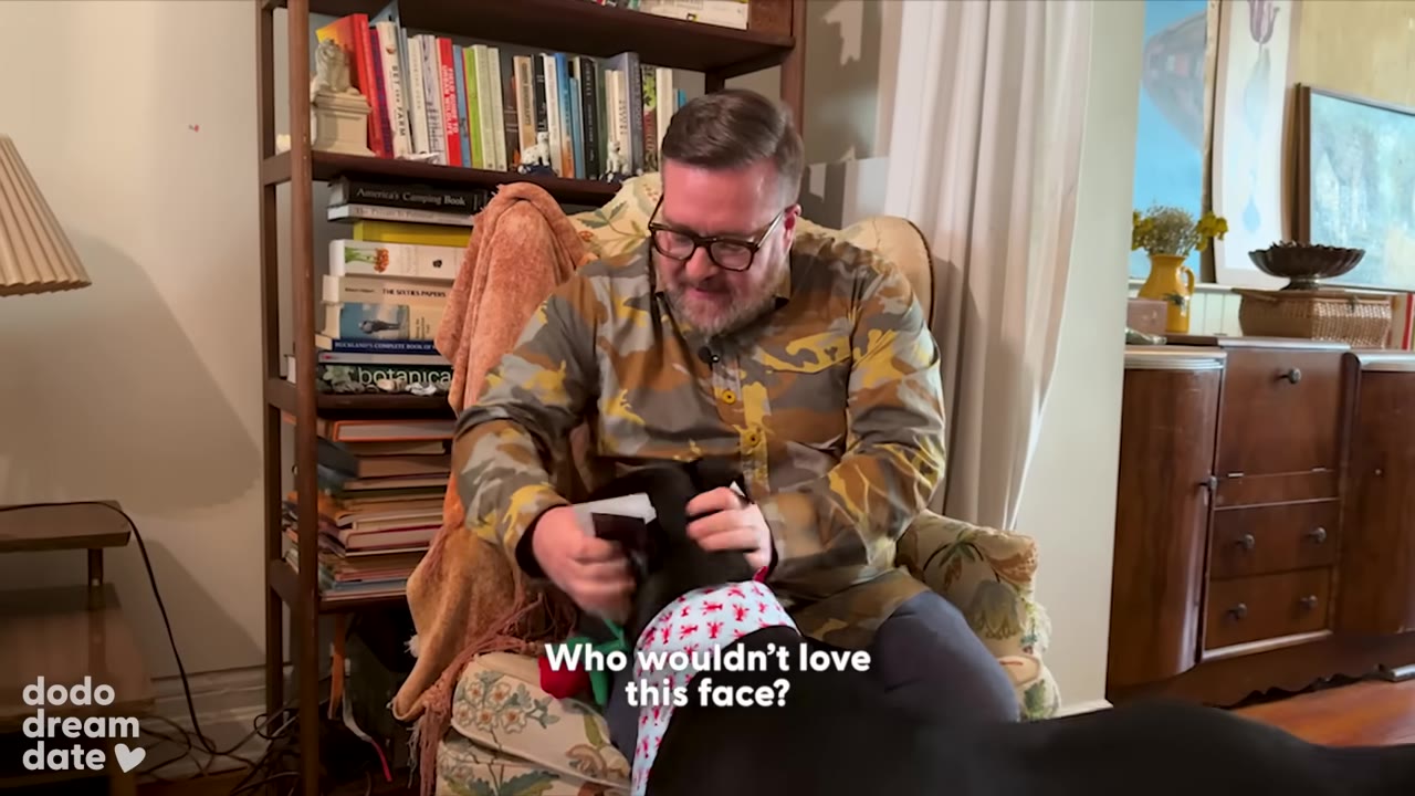 A man in a camouflage shirt and glasses sits in an armchair, affectionately cradling the face of a black dog wearing a patterned bandana.