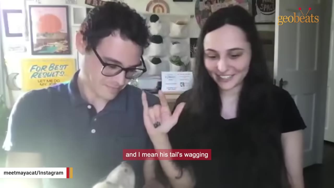 A man wearing glasses makes the sign language gesture for 'I love you' with his hand while looking down at a dog, next to a smiling woman.
