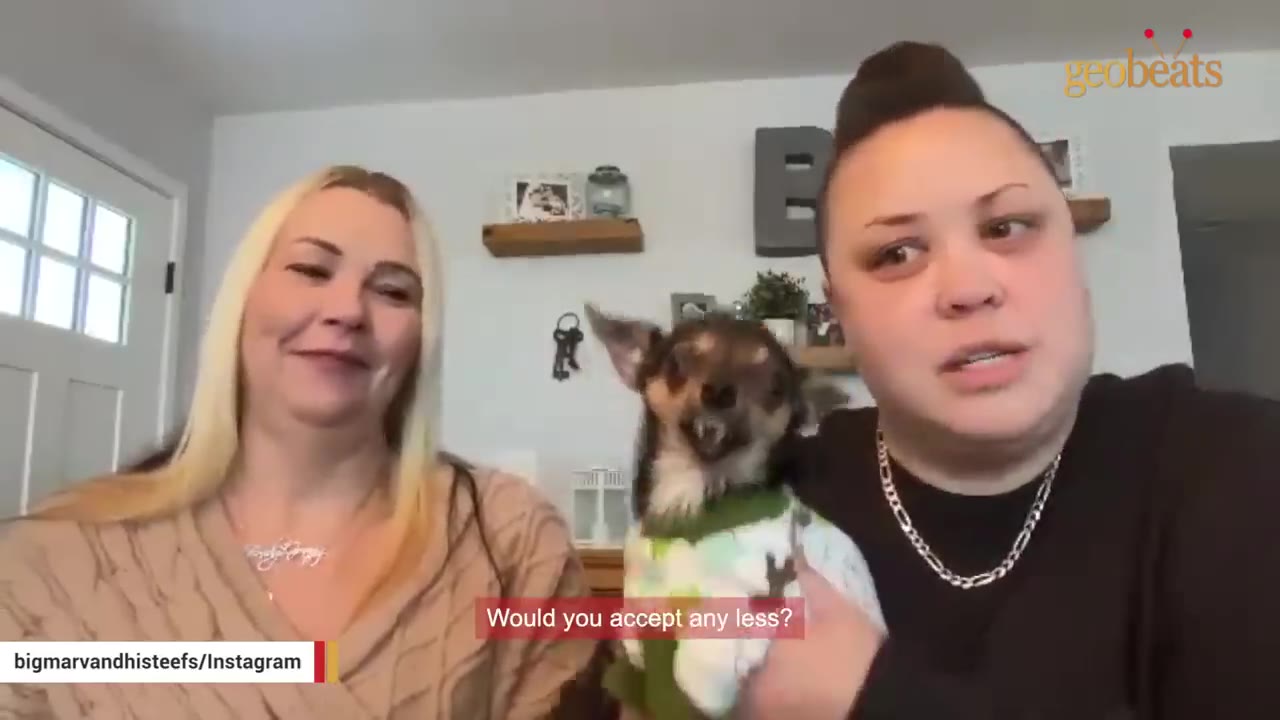 Two women, one blonde and one with dark hair styled up, smile and hold a small chihuahua wearing a green and white sweater.