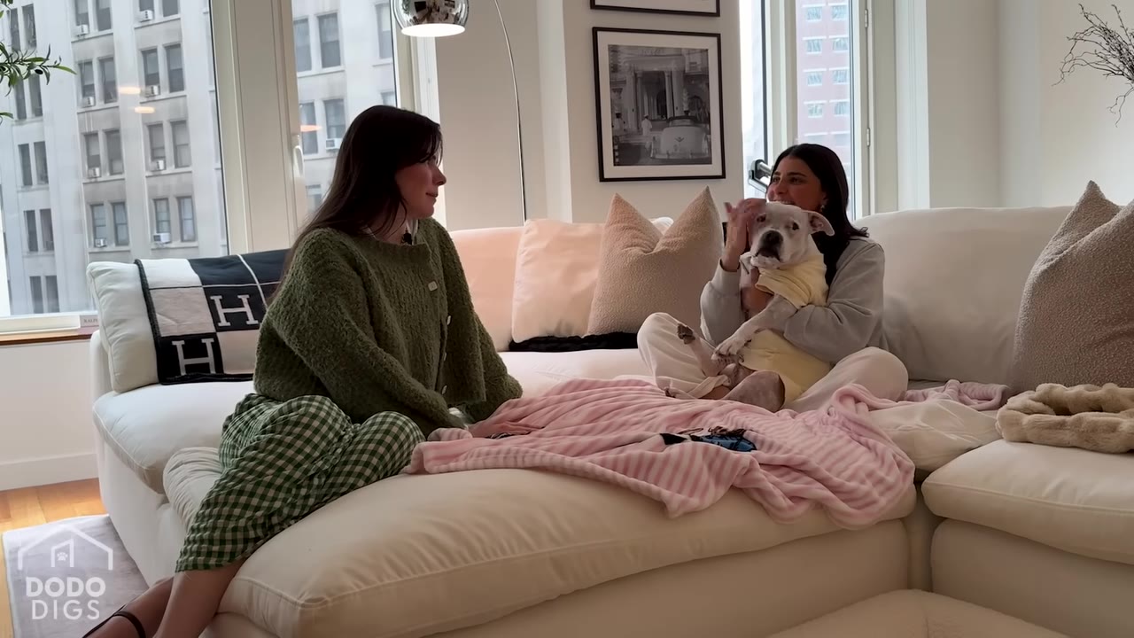 Two women sit on a white sectional sofa in a bright apartment, with one woman holding a white dog wearing a yellow shirt.
