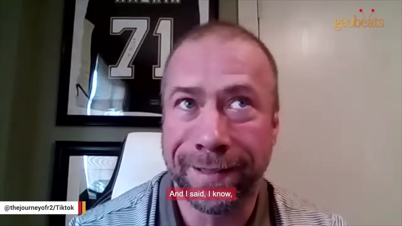 A close-up of a man with short hair and a beard speaking to the camera, with framed sports jerseys visible in the background.